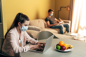 selective focus of freelancer in medical mask using laptop near man with digital tablet at home