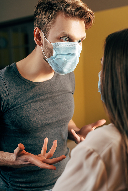 selective focus of emotional man in medical mask gesturing while quarreling with girlfriend at home