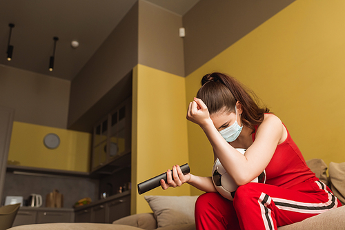 upset sportswoman in medical mask holding remote controller near football while watching championship in living room