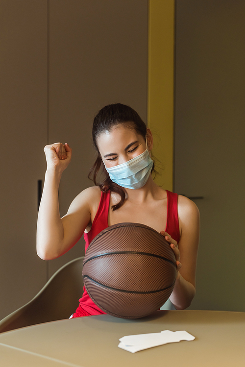 sportive woman in medical mask and closed eyes cheering near basketball