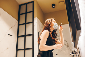 low angle view of woman in medical mask and black dress holding cosmetic brush with eye shadow in bathroom
