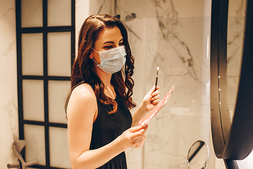 woman in medical mask and black dress holding night party tickets and cosmetic brush, end of quarantine concept