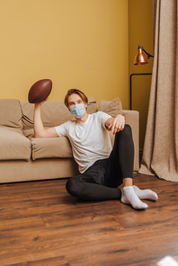 young man in medical mask holding american football while sitting on floor in living room