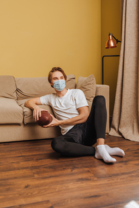 young man in medical mask holding american football and sitting on floor in living room