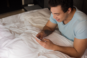 smiling young mixed race man using smartphone in bed on quarantine