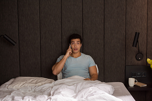handsome surprised mixed race man talking on smartphone in bed on quarantine