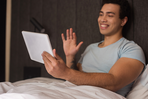 happy mixed race man waving while having video chat on digital tablet in bed during self isolation
