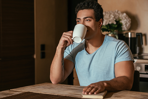 mixed race man drinking coffee and holding book on kitchen during self isolation