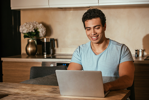 smiling mixed race freelancer working with laptop on kitchen during self isolation