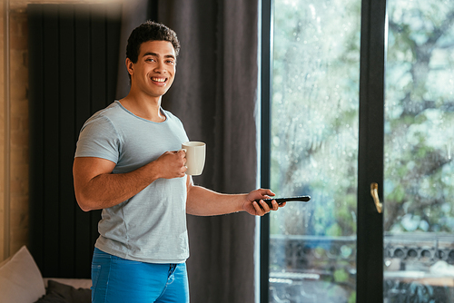 smiling mixed race man holding cup of coffee and remote controller at home during quarantine