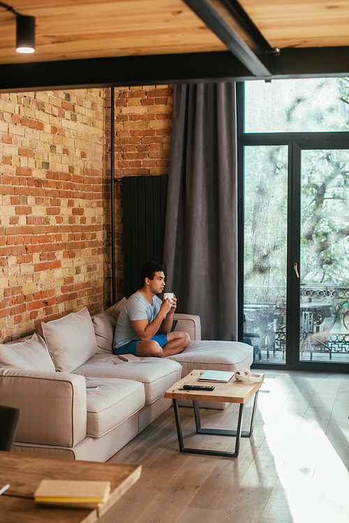 handsome mixed race man holding cup of coffee on sofa in living room during quarantine