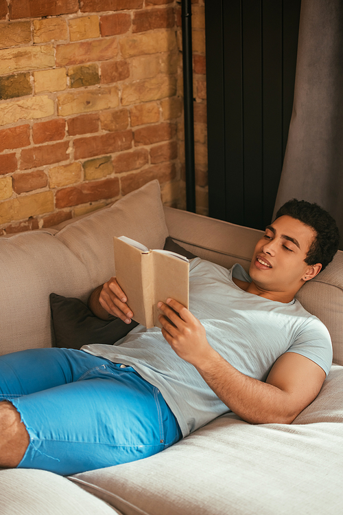 handsome mixed race man reading book while chilling on sofa during self isolation