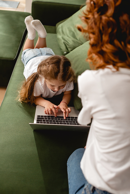 back view of curly mother near daughter using laptop in living room
