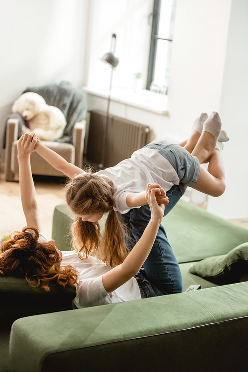 curly mother lying on sofa, lifting daughter and holding hands in living room