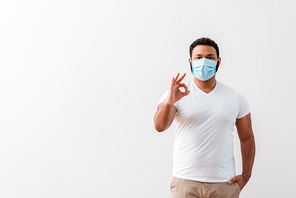 african american man in medical mask standing with hand in pocket and showing ok sign near white wall