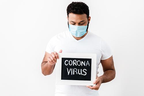 african american man in medical mask looking at chalkboard with coronavirus lettering near white wall