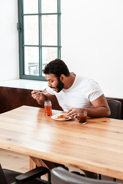 african american man holding spoon with sweet jam near opened mouth and toast bread on plate and cup of coffee