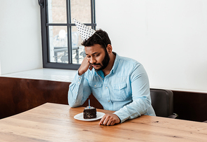 sad african american man in party cap celebrating birthday alone and looking at birthday cake