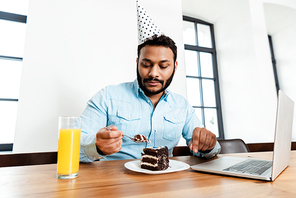 african american man in party cap looking at tasty birthday cake near laptop and glass of orange juice