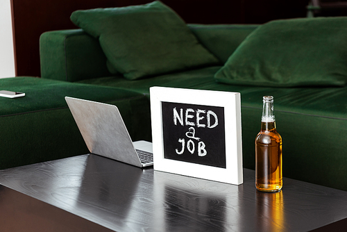 laptop near chalkboard with need a job lettering and bottle of beer near sofa