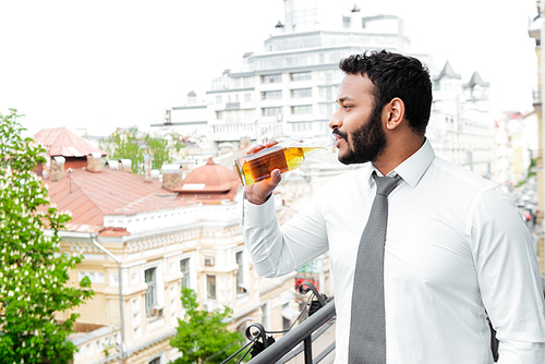 side view of bearded african american man in suit drinking beer on balcony