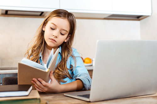 selective focus of cute kid holding book near laptop while online studying at home