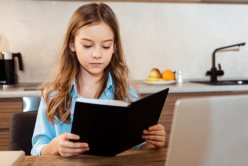 selective focus of cute kid reading book near laptop while e-learning at home