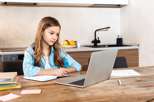 selective focus of cute kid writing in notebook near laptop while e-learning at home