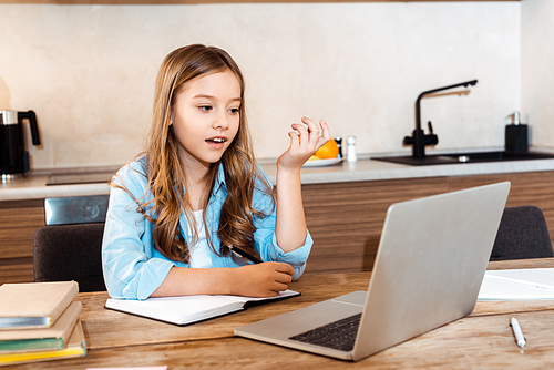 selective focus of surprised kid writing in notebook near laptop while e-learning at home