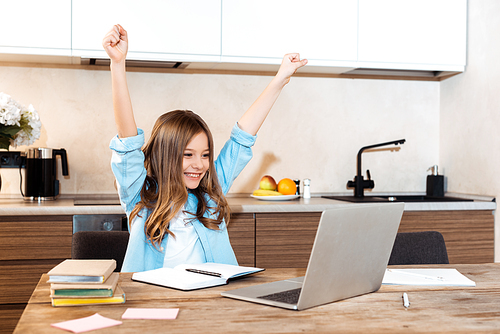 selective focus of happy kid with hands above head looking at laptop while e-learning at home