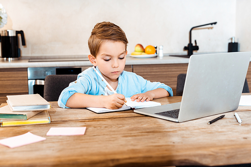 selective focus of boy writing in notebook near laptop while e-learning at home