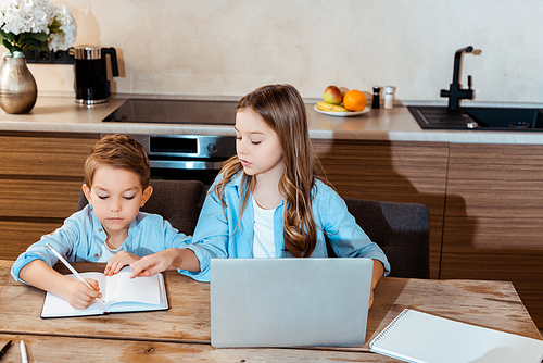 sister looking at brother writing in notebook near laptop while e-learning at home