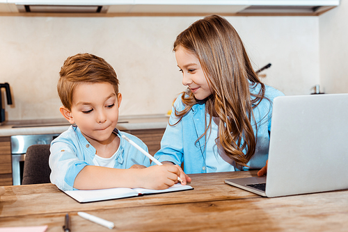 selective focus of happy sister looking at brother writing in notebook near laptop while e-learning at home