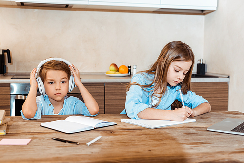 kid writing in notebook near brother in wireless headphones at home