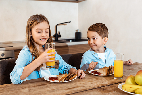 selective focus of happy child holding glass of orange juice near brother and tasty breakfast