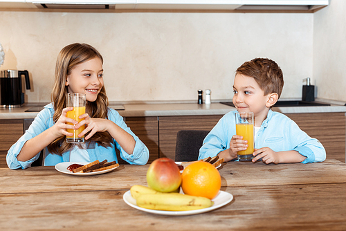 selective focus of happy siblings looking at each other and holding glasses of orange juice near tasty breakfast