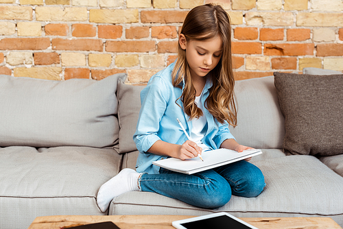 kid sitting on sofa and writing in notebook near digital tablet with blank screen