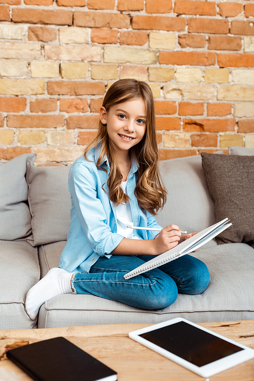 happy kid sitting on sofa and writing in notebook near digital tablet with blank screen