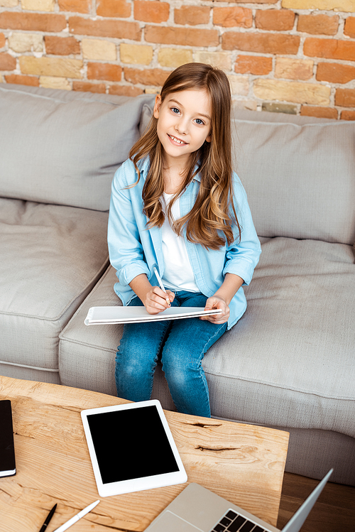 cheerful kid sitting on sofa and writing in notebook near digital tablet with blank screen