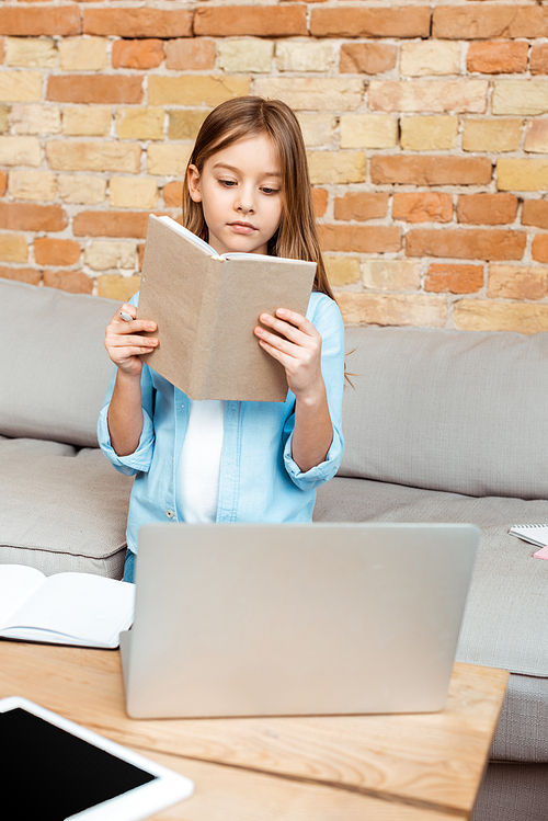 selective focus of cute kid reading book near laptop and digital tablet