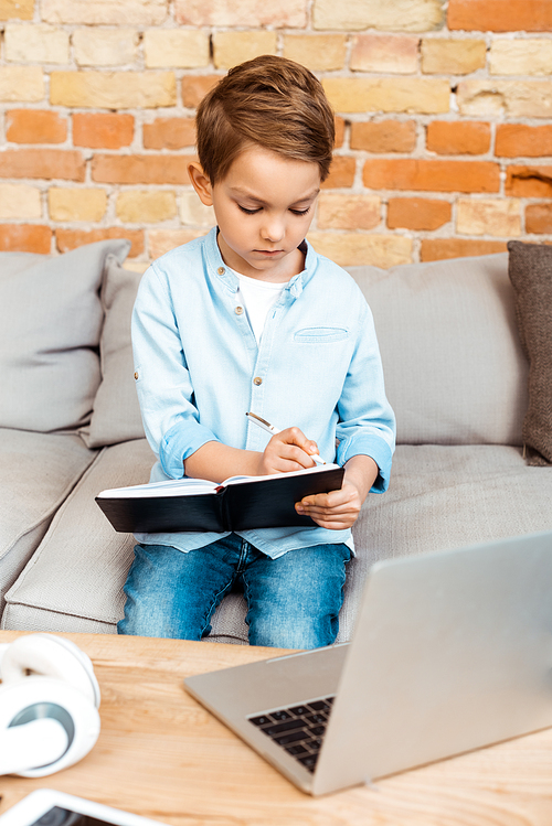 boy writing in notebook near laptop while e-learning at home