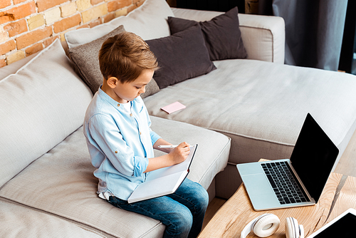 adorable boy writing in notebook near gadgets with blank screen while e-learning at home