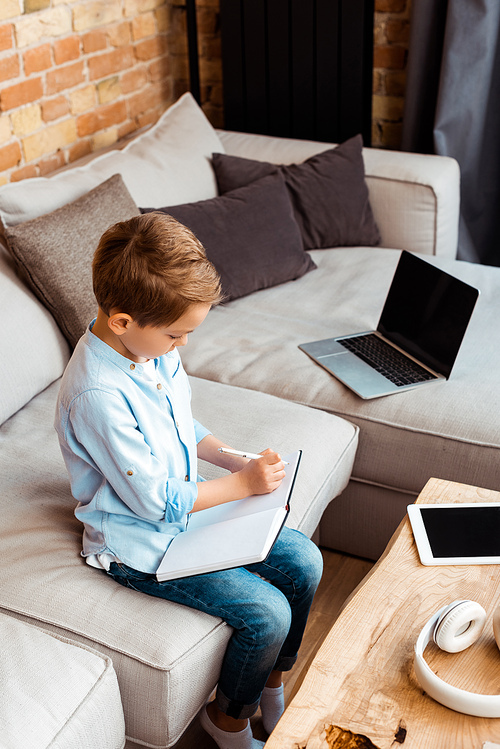 cute boy writing in notebook near gadgets with blank screen while e-learning at home