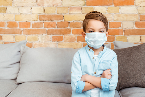 boy in medical mask with crossed arms near brick wall in living room