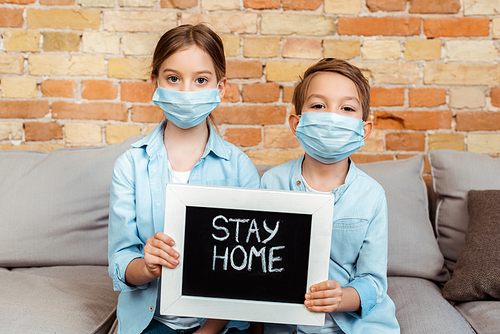 siblings in medical masks holding chalkboard with stay home lettering