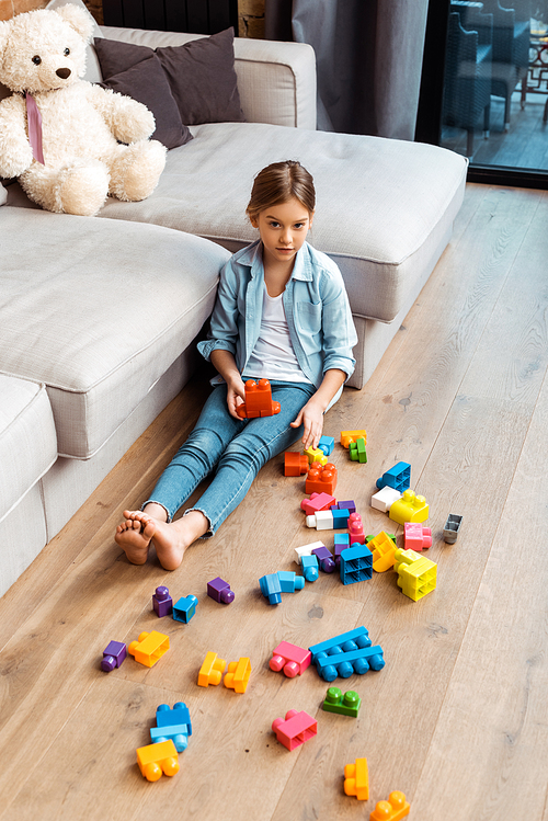 cute kid sitting on floor near building blocks and sofa