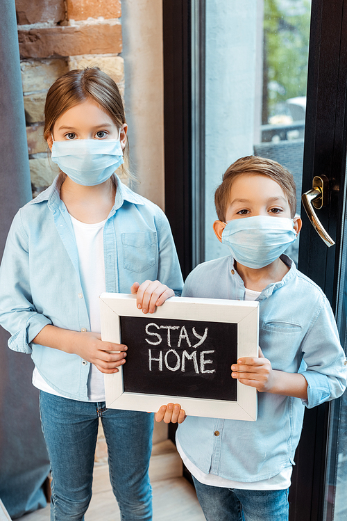 siblings in medical masks holding chalkboard with stay home lettering