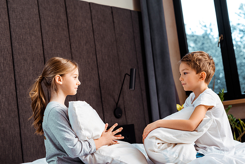 side view of sister holding pillow and looking at brother in bedroom