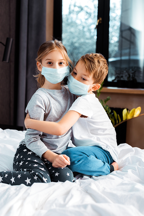 cute brother hugging sister in medical mask while sitting on bed