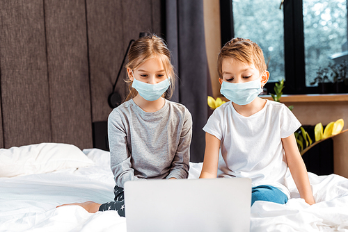 selective focus of kids in medical masks looking at laptop while e-learning in bedroom